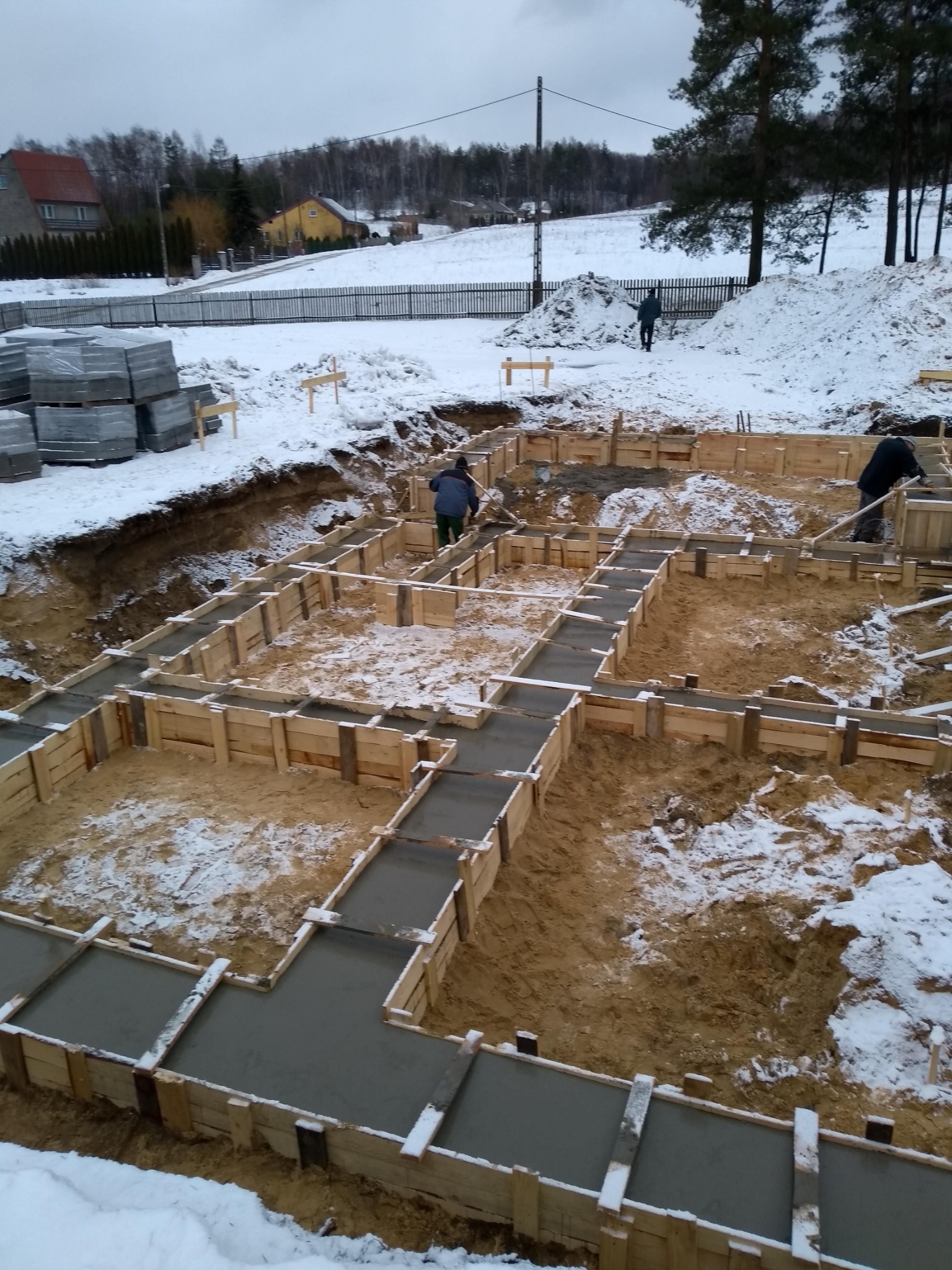Zimowy krajobraz budowy fundamentów: drewniane szalunki wypełnione świeżym betonem, częściowo pokryte śniegiem, widoczni pracownicy w oddali, w tle ośnieżone domy i drzewa.