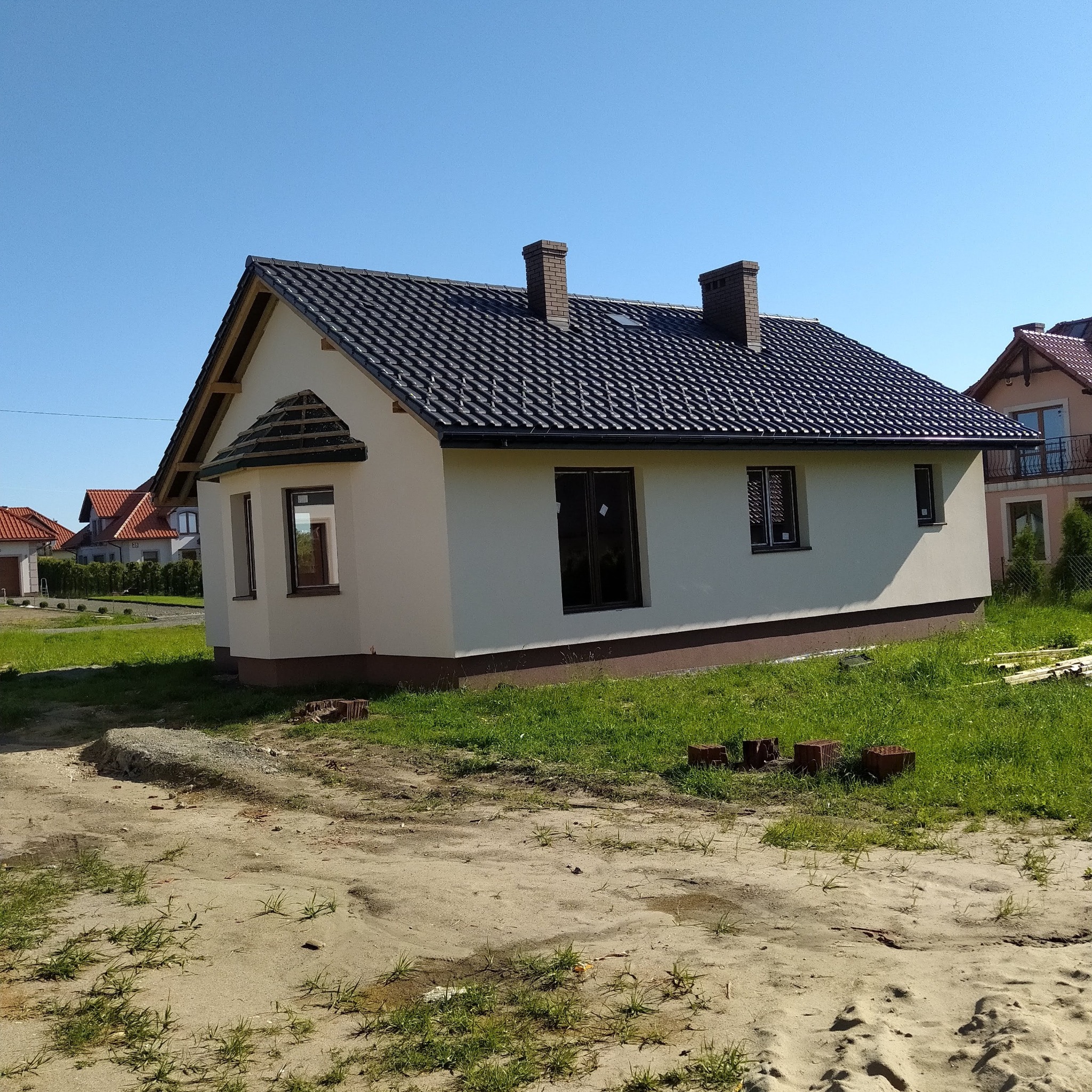 Niedokończony dom jednorodzinny z ciemną dachówką, widoczne cegły na trawie i niezabezpieczone okna, w tle inne domy.