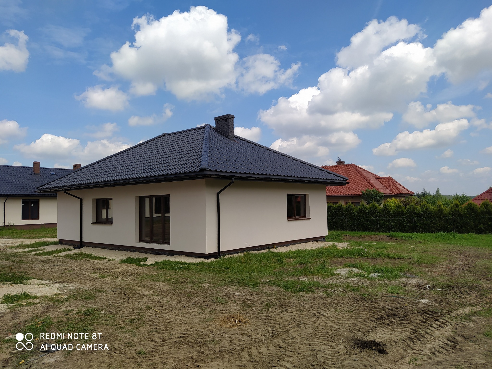 Nowoczesny dom murowany w Ryczowie z ciemną dachówką, jasną elewacją i widocznym kominem, otoczony świeżo przygotowanym terenem pod trawnik.