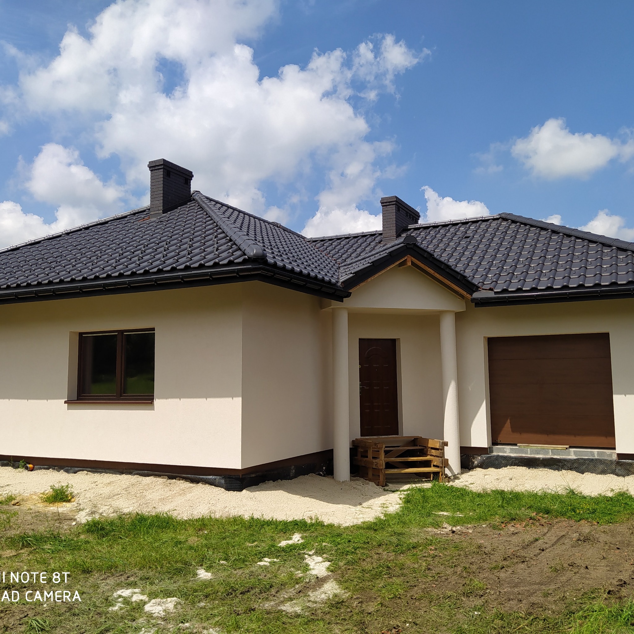 Nowoczesny parterowy dom z ciemną dachówką, kremową elewacją i brązową stolarką okienną oraz garażową, widok z zewnątrz, na trawniku piasek.