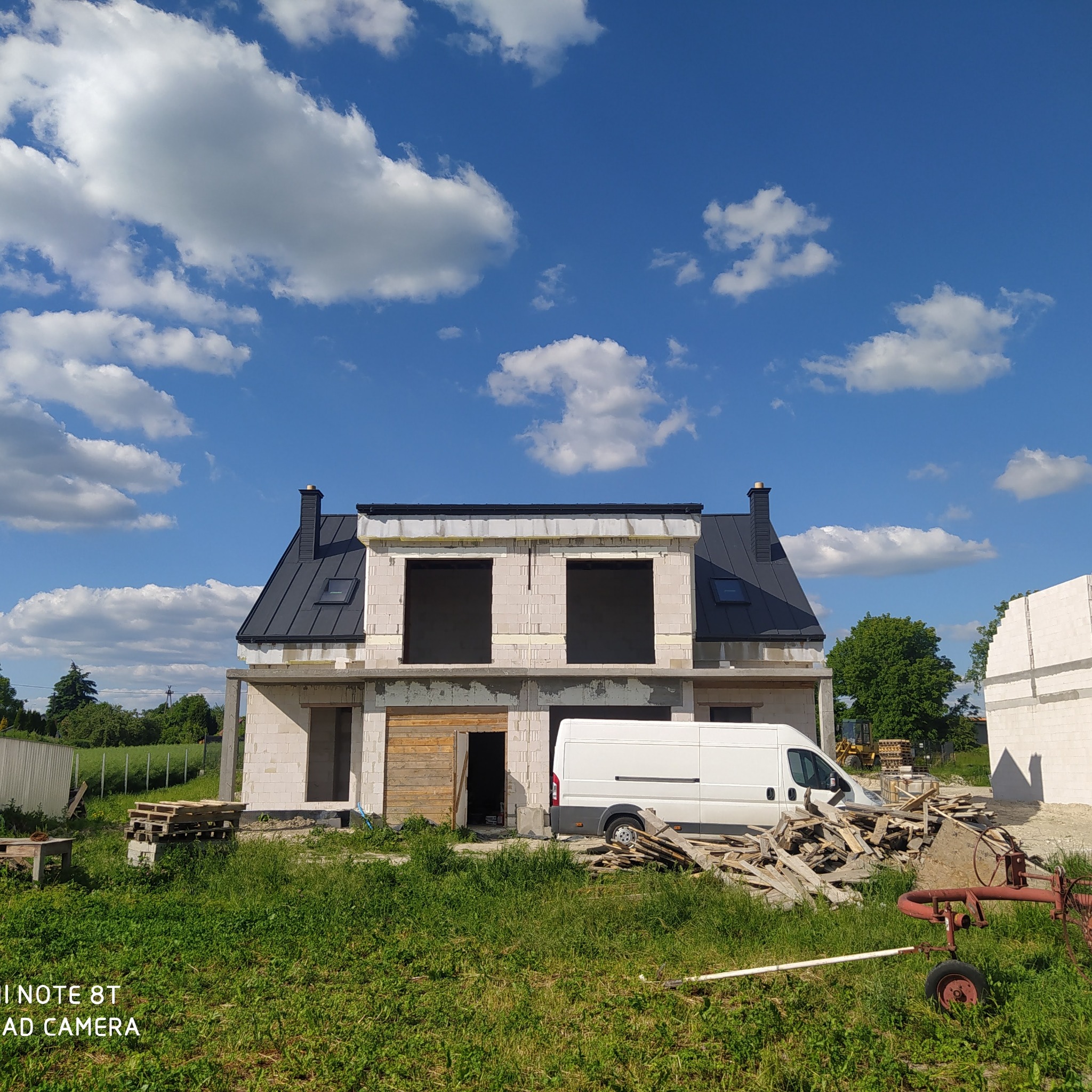 Niedokończony dwukondygnacyjny budynek z pustaków z czarnym dachem i kominami, biały van na tle, rozrzucone deski i maszyna rolnicza na trawiastym terenie w słoneczny dzień.