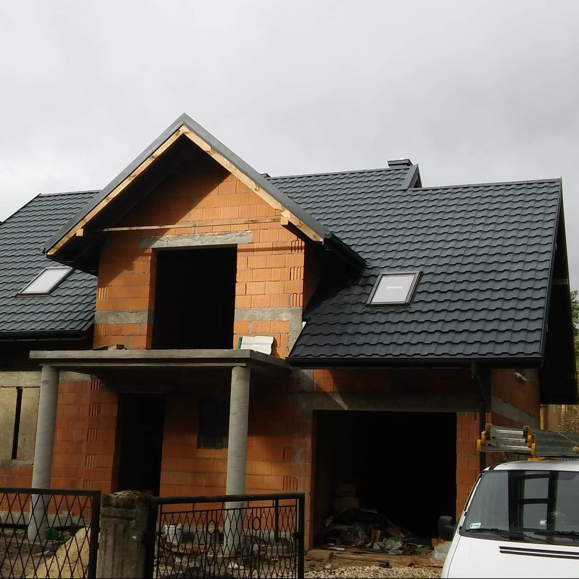 Niedokończony dom z czerwonej cegły z zamontowaną czarną dachówką, widoczne okno dachowe, stan surowy otwarty, podjazd z gruzem, samochód dostawczy w tle.