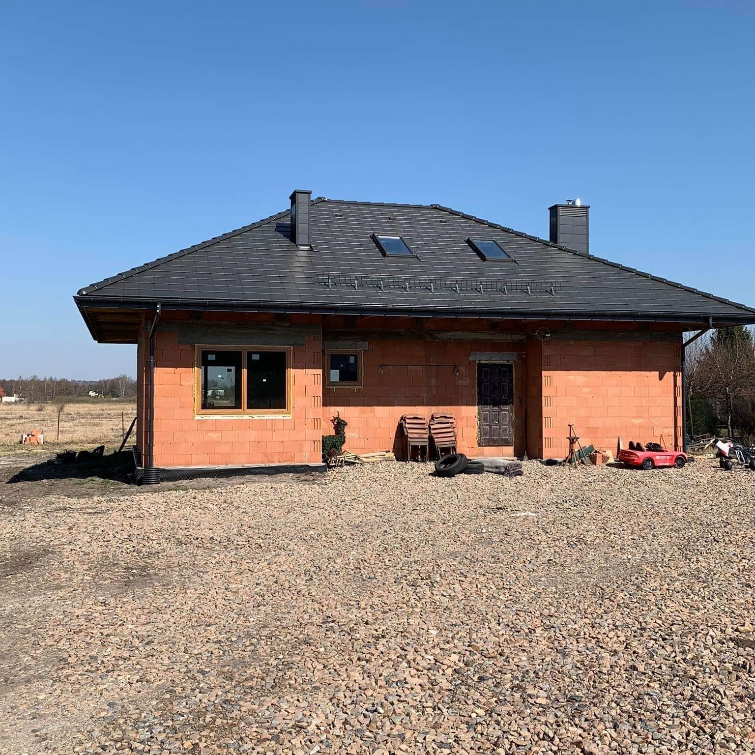 Surowy stan domu jednorodzinnego z czerwonej cegły, czarnym dachem z dachówkami i trzema oknami dachowymi, na żwirowym podłożu w słoneczny dzień.