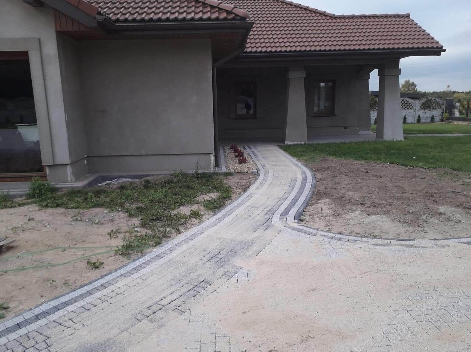 Ułożona kostka brukowa tworząca ścieżkę do domu z kolumnami, z widocznym fragmentem trawnika i rabatą kwiatową. Jasna kostka z ciemnymi obrzeżami.