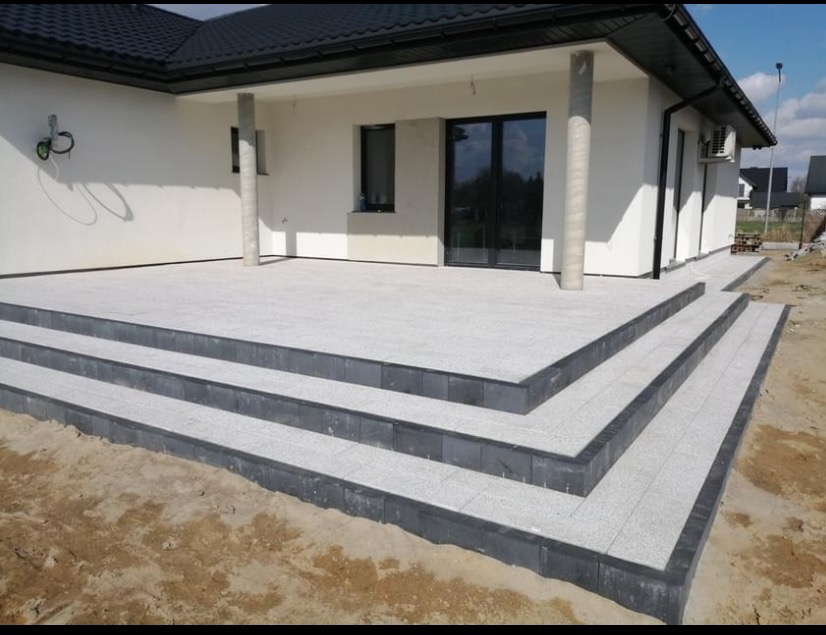 Jasnoszara kostka brukowa na tarasie z trzema stopniami, obrzeża z ciemnej kostki. Biały dom z kolumnami w tle, częściowo widoczne niezagospodarowane podwórko.