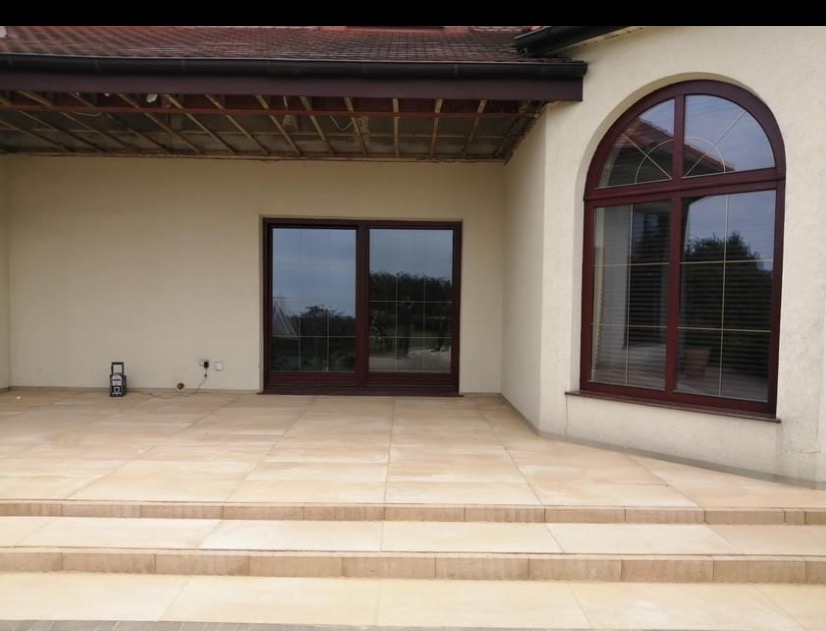 Jasna, elegancka kostka brukowa na tarasie przed domem z dużym oknem łukowym i przesuwnymi drzwiami tarasowymi. Widoczne schody z kostki.