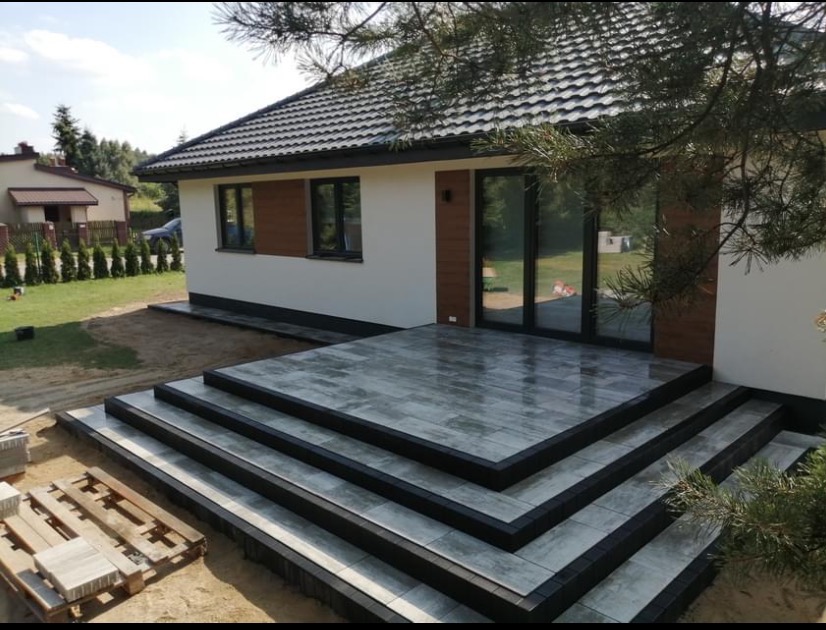 Nowoczesny taras z szarej kostki brukowej z czarnymi obrzeżami, prowadzący do domu z dużymi oknami, widok z góry, częściowo ułożona kostka na palecie.