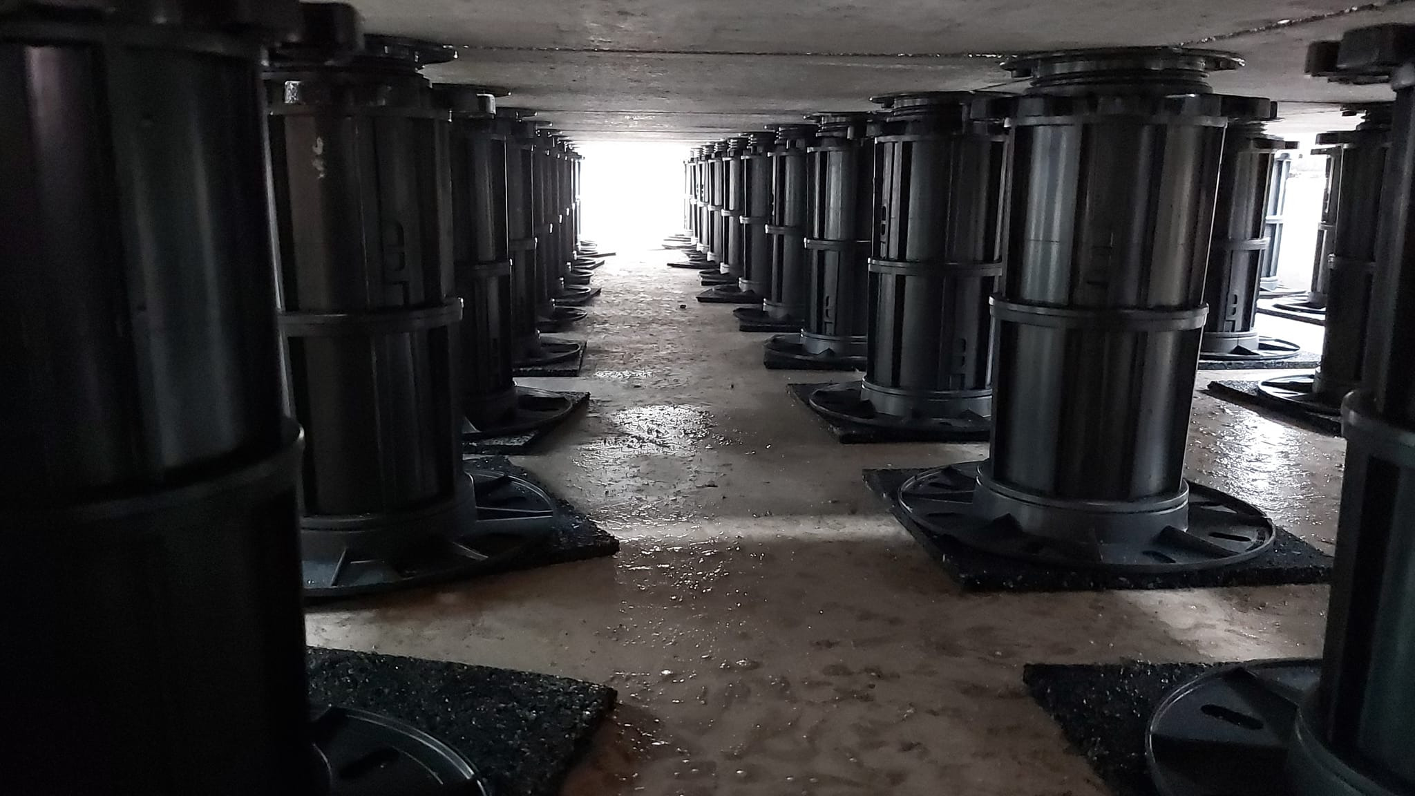 Równomiernie rozmieszczone, czarne, regulowane wsporniki pod taras betonowy, widoczne od spodu, z perspektywą zbiegającą się w kierunku jasnego światła na końcu tunelu