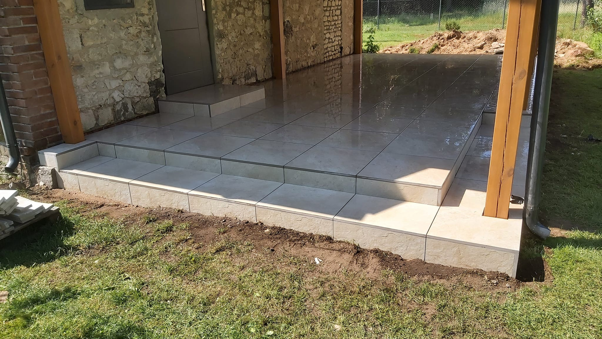 Wyłożony jasnymi płytkami taras z dwoma stopniami, widoczne drewniane słupy konstrukcyjne i kamienne ściany budynku.