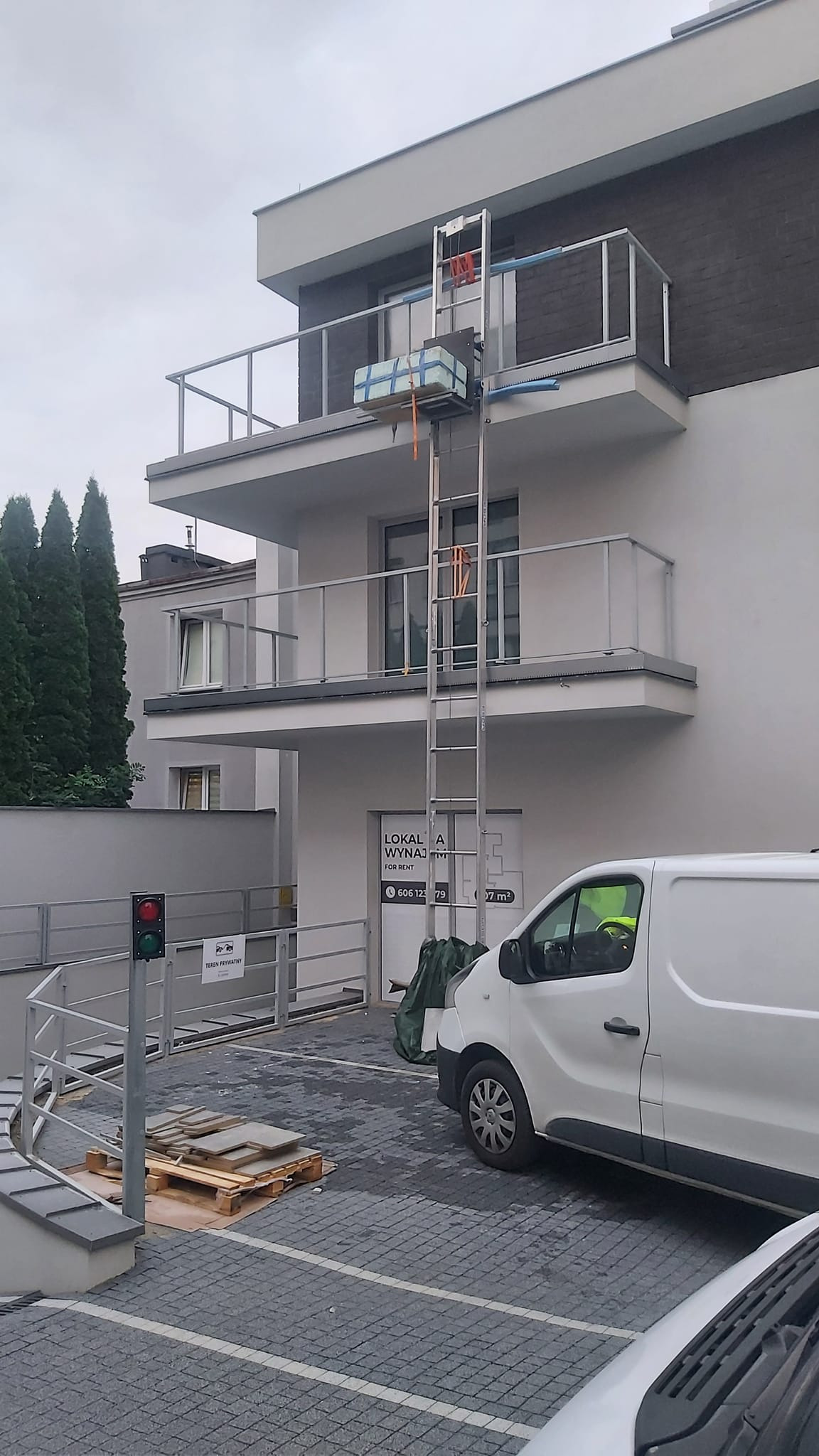 Winda meblowa transportuje paczkę na balkon nowoczesnego budynku z szarą elewacją i szklanymi balustradami; na dole zaparkowany biały samochód dostawczy.