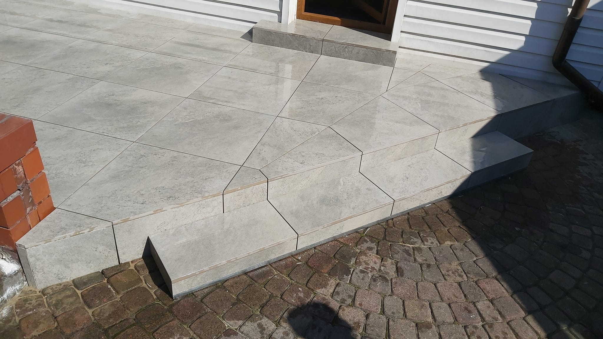 Wykończone schody wejściowe do domu, pokryte szarymi płytkami ceramicznymi o nieregularnym wzorze, z widocznym fragmentem elewacji z sidingu i kostki brukowej.