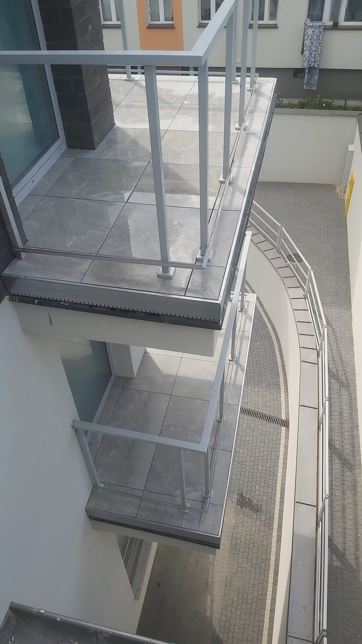 Widok z góry na balkony wykończone szarymi płytkami imitującymi marmur, z białymi balustradami, widoczne z góry na dziedziniec wyłożony kostką brukową.