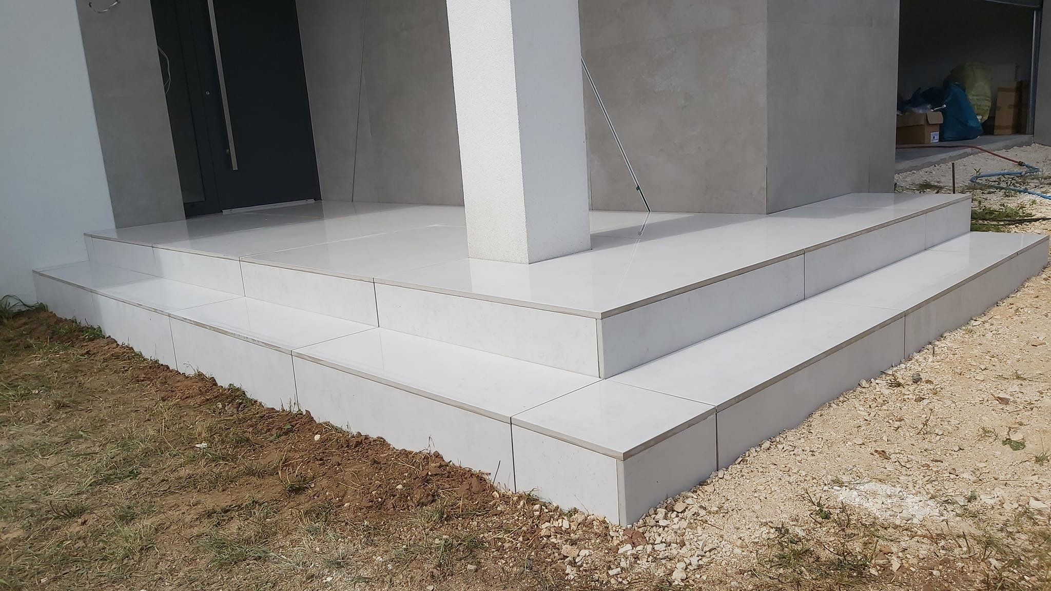 Wyłożone białymi płytkami schody wejściowe do domu z kolumną, widoczne niedoskonałości w ułożeniu na krawędziach stopni.