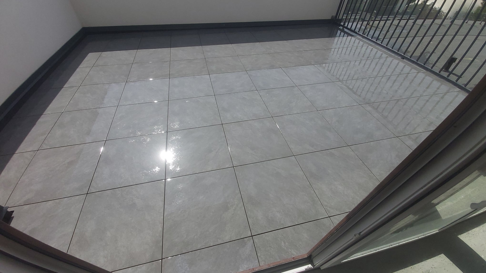 Balkon wyłożony szarymi, kwadratowymi płytkami gresowymi, widoczne odbicie światła na powierzchni płytek, barierka balkonowa w tle.