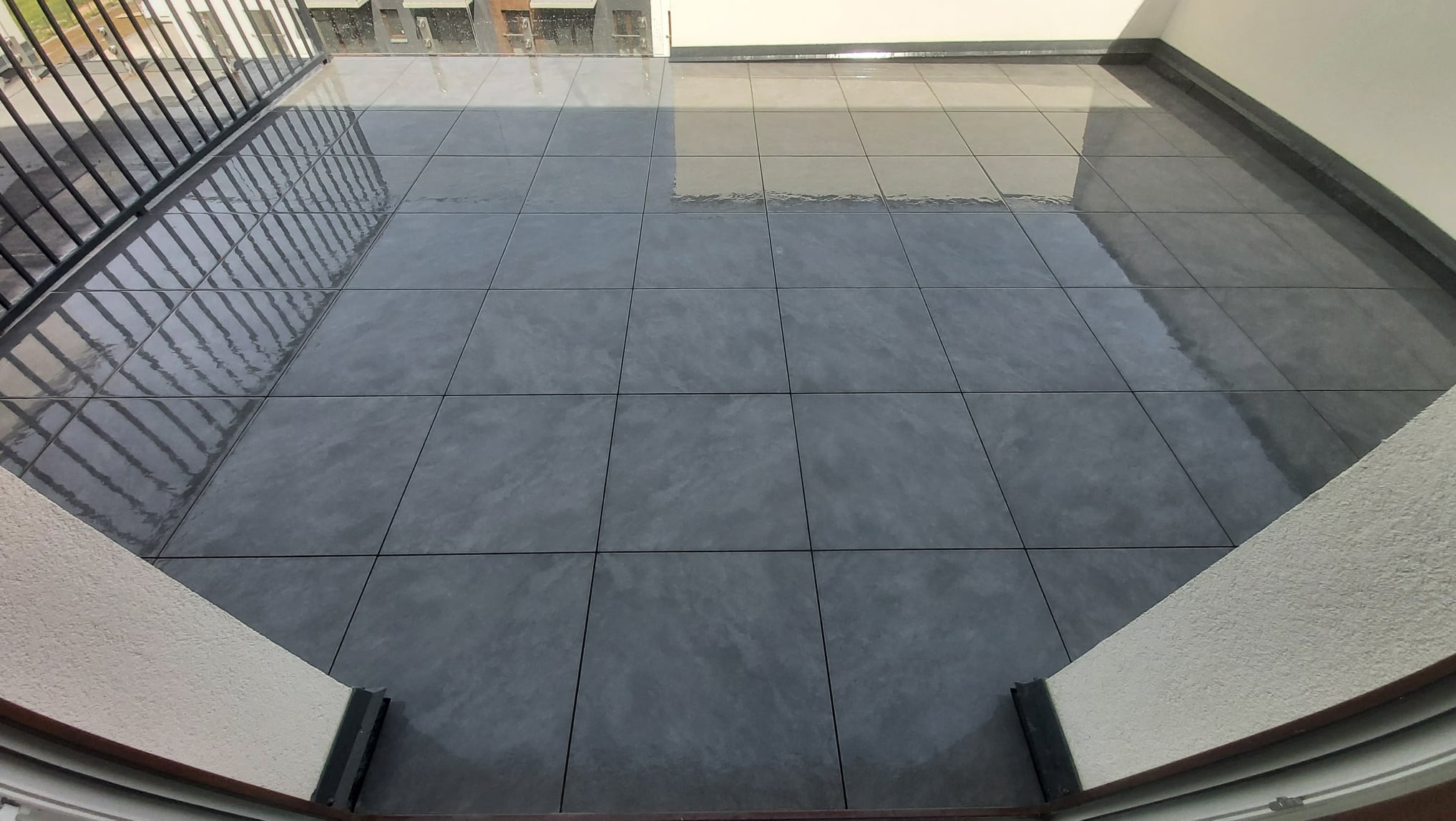 Balkon z szarymi, kwadratowymi płytkami podłogowymi, widoczne refleksy światła na powierzchni płytek, czarna balustrada w tle.