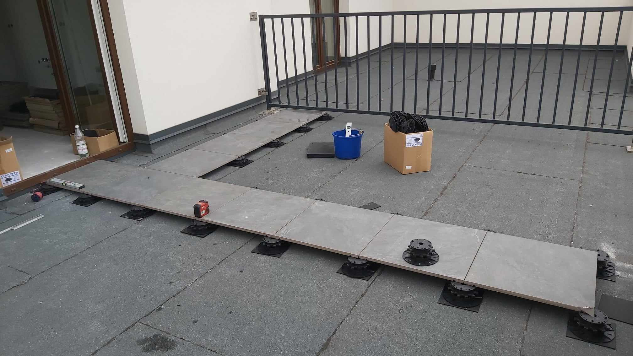 Montaż szarych płyt tarasowych na regulowanych wspornikach na balkonie, widoczne narzędzia pracy, poziomica i laserowy dalmierz, w tle barierka balkonowa i drzwi balkonowe.