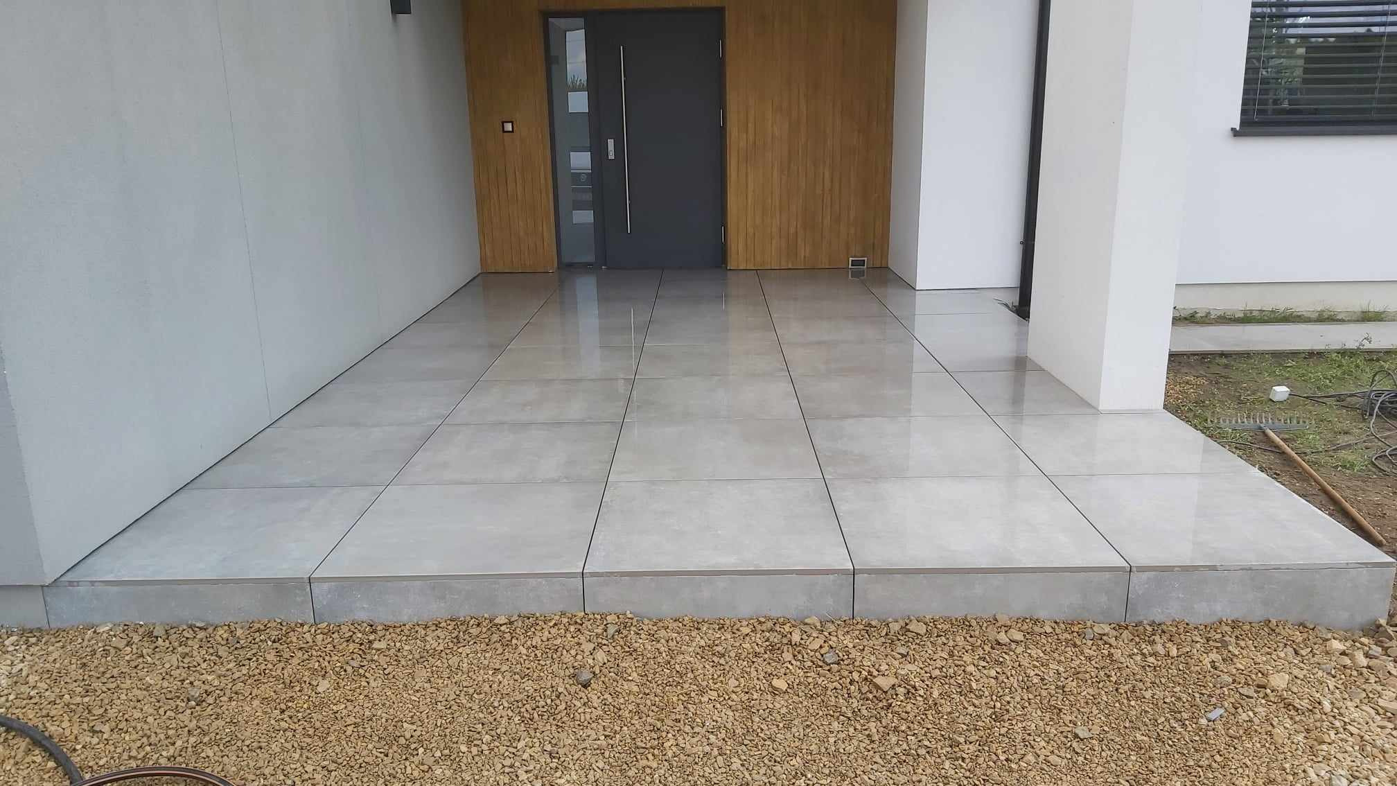 Nowoczesny taras przed wejściem do domu wyłożony dużymi, jasnoszarymi płytkami gresowymi z widocznym progiem i drobnym żwirem na obrzeżach.