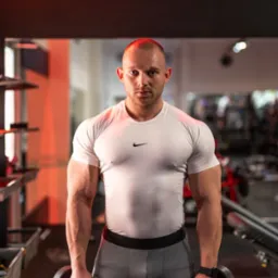 Mężczyzna z ogoloną głową, w sportowym stroju i z hantlami 12,5 kg w dłoniach, pozuje w siłowni oświetlonej czerwonym światłem.