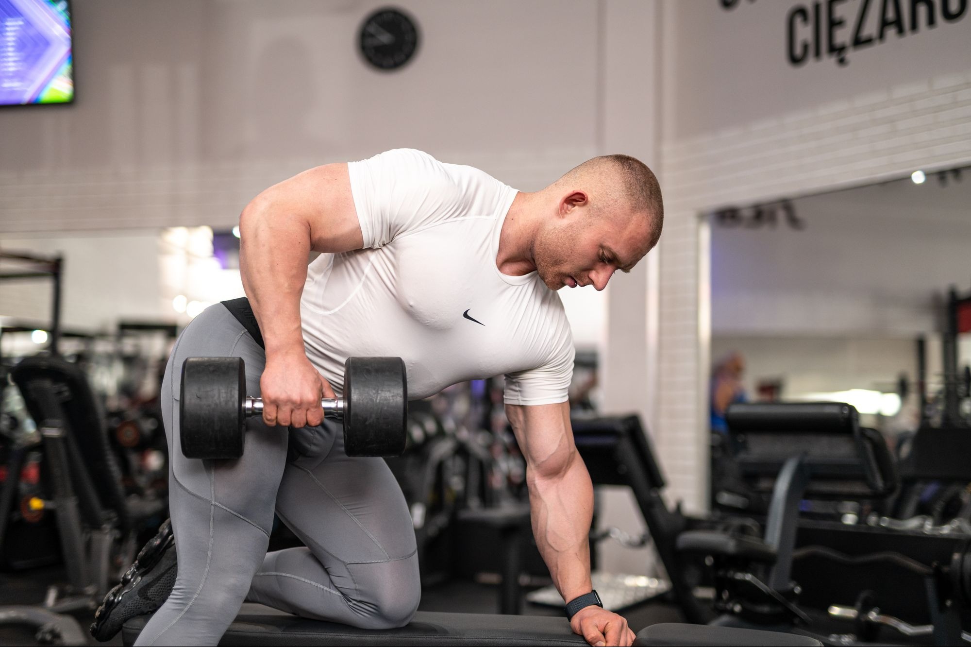 Mężczyzna w sportowym stroju, klęcząc na ławce w siłowni, wykonuje ćwiczenie wiosłowania hantlą, skupiony wzrok, widoczne napięcie mięśni ramion i pleców.
