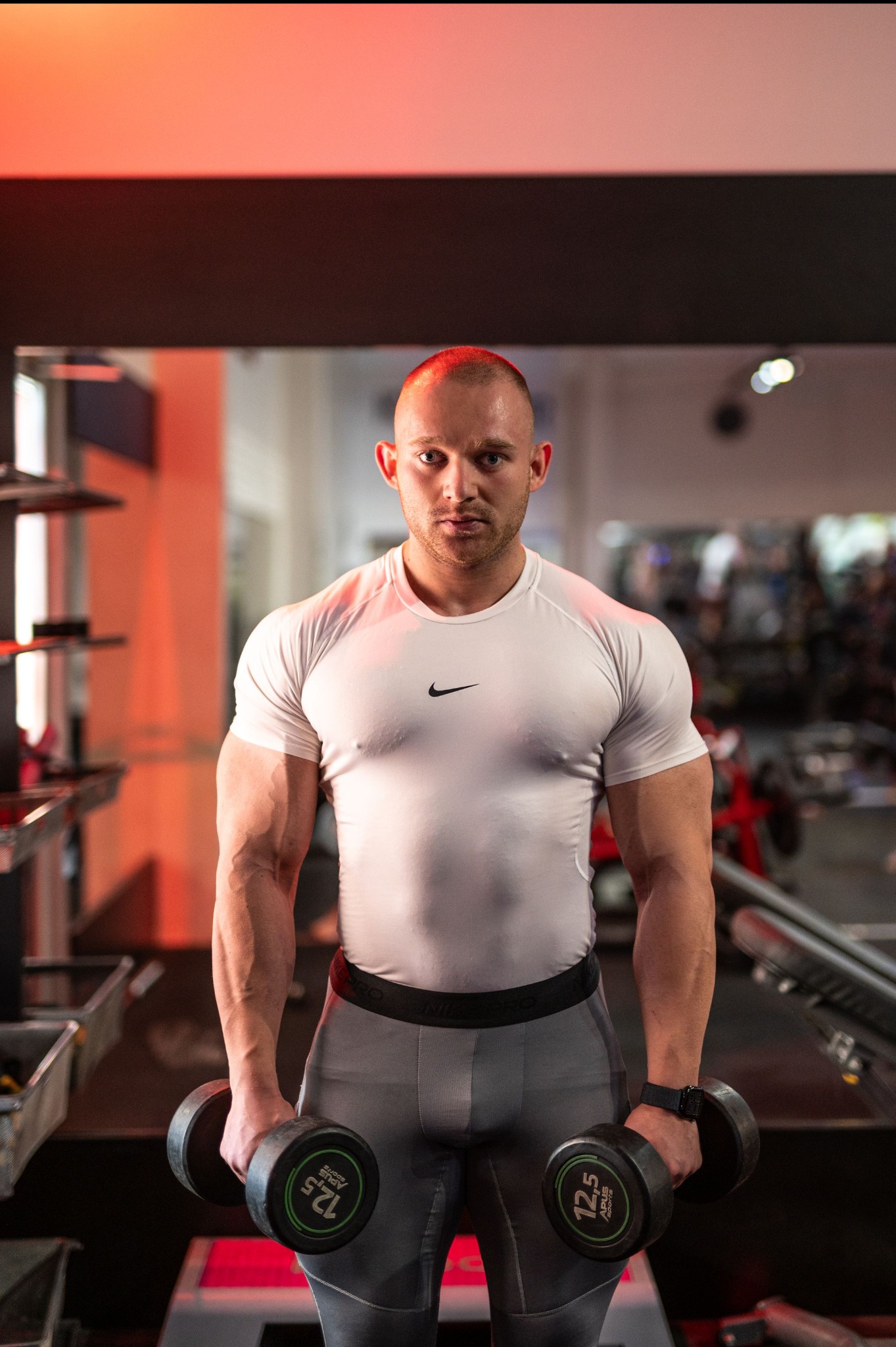 Mężczyzna z ogoloną głową, w sportowym stroju i z hantlami 12,5 kg w dłoniach, pozuje w siłowni oświetlonej czerwonym światłem.