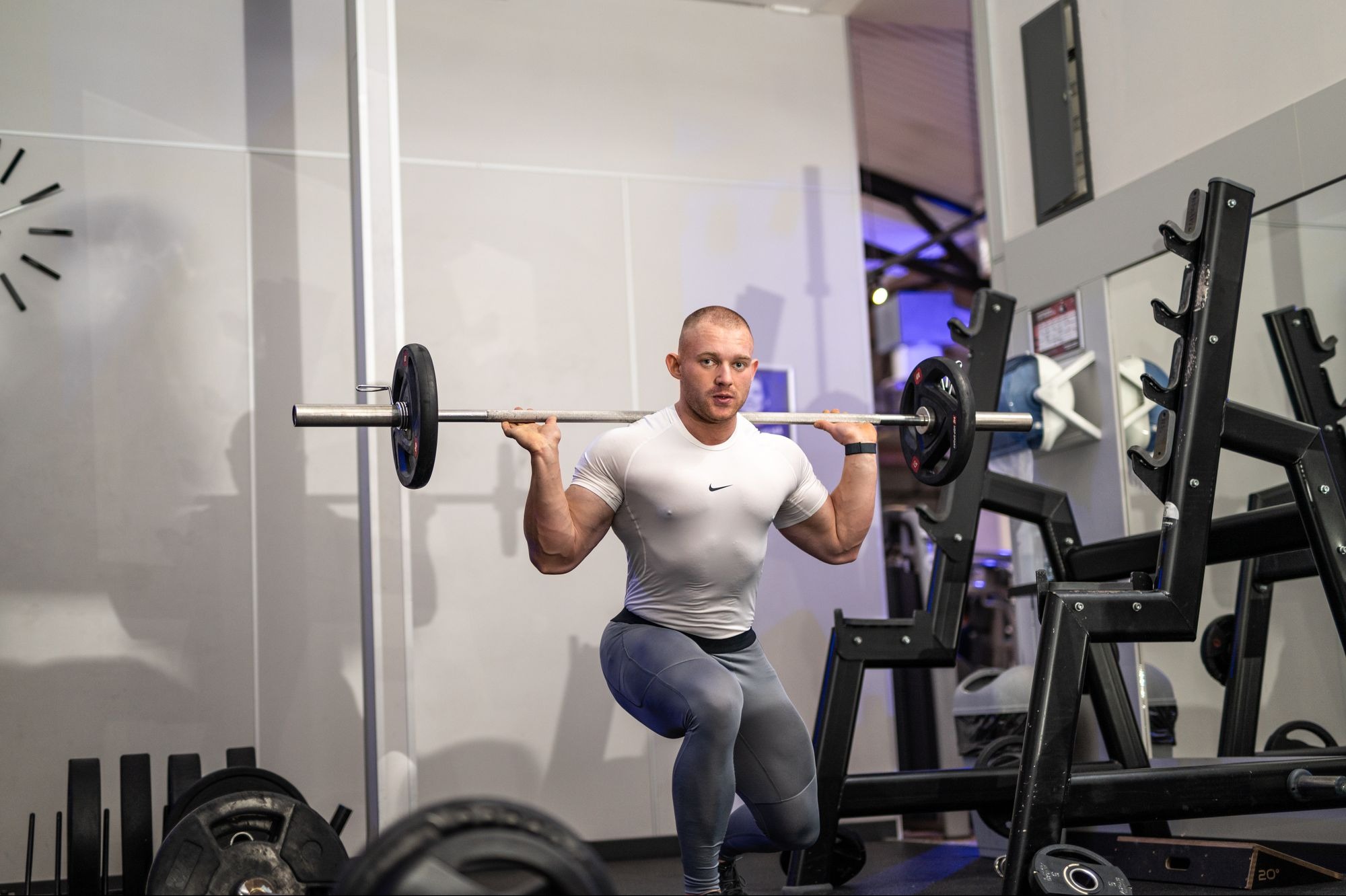 Mężczyzna w białej koszulce i szarych legginsach wykonuje przysiad ze sztangą na barkach w siłowni, widoczny stojak na sztangę i zegar na ścianie w tle.