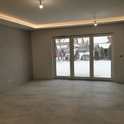 P.K Projekt Przemysław Kostecki - Pusty pokój z szarymi płytkami podłogowymi, panelami ściennymi i oknami balkonowymi z widokiem na zimowy krajobraz; widoczne puszki z farbą w rogu.