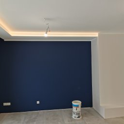 P.K Projekt Przemysław Kostecki - Pomieszczenie po malowaniu: granatowa ściana, biały narożnik, szara podłoga z płytek, wiadro po farbie przy ścianie, podsufitowe oświetlenie LED, surowa żarówka.