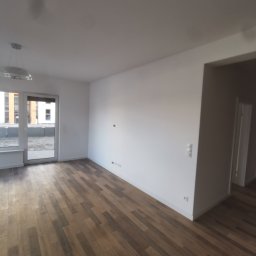 P.K Projekt Przemysław Kostecki - Puste, jasne pomieszczenie z panelami podłogowymi imitującymi drewno ułożonymi wzdłuż, białe ściany, okno balkonowe z widokiem na inne budynki, widoczny sufit z lampą.