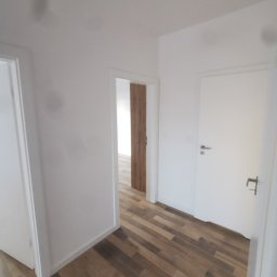 P.K Projekt Przemysław Kostecki - Korytarz po remoncie z drewnopodobną podłogą ułożoną w jodełkę, białe ściany i drzwi, widok na kolejne pomieszczenie z drewnianą ścianą.