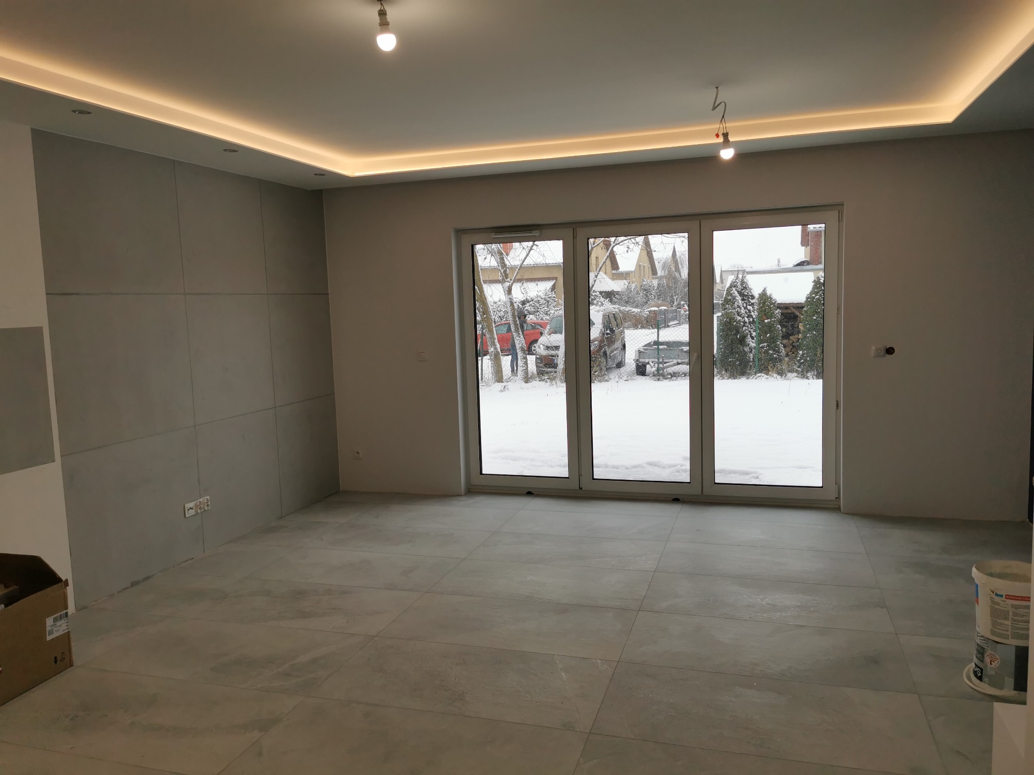 Pusty pokój z szarymi płytkami podłogowymi, panelami ściennymi i oknami balkonowymi z widokiem na zimowy krajobraz; widoczne puszki z farbą w rogu.