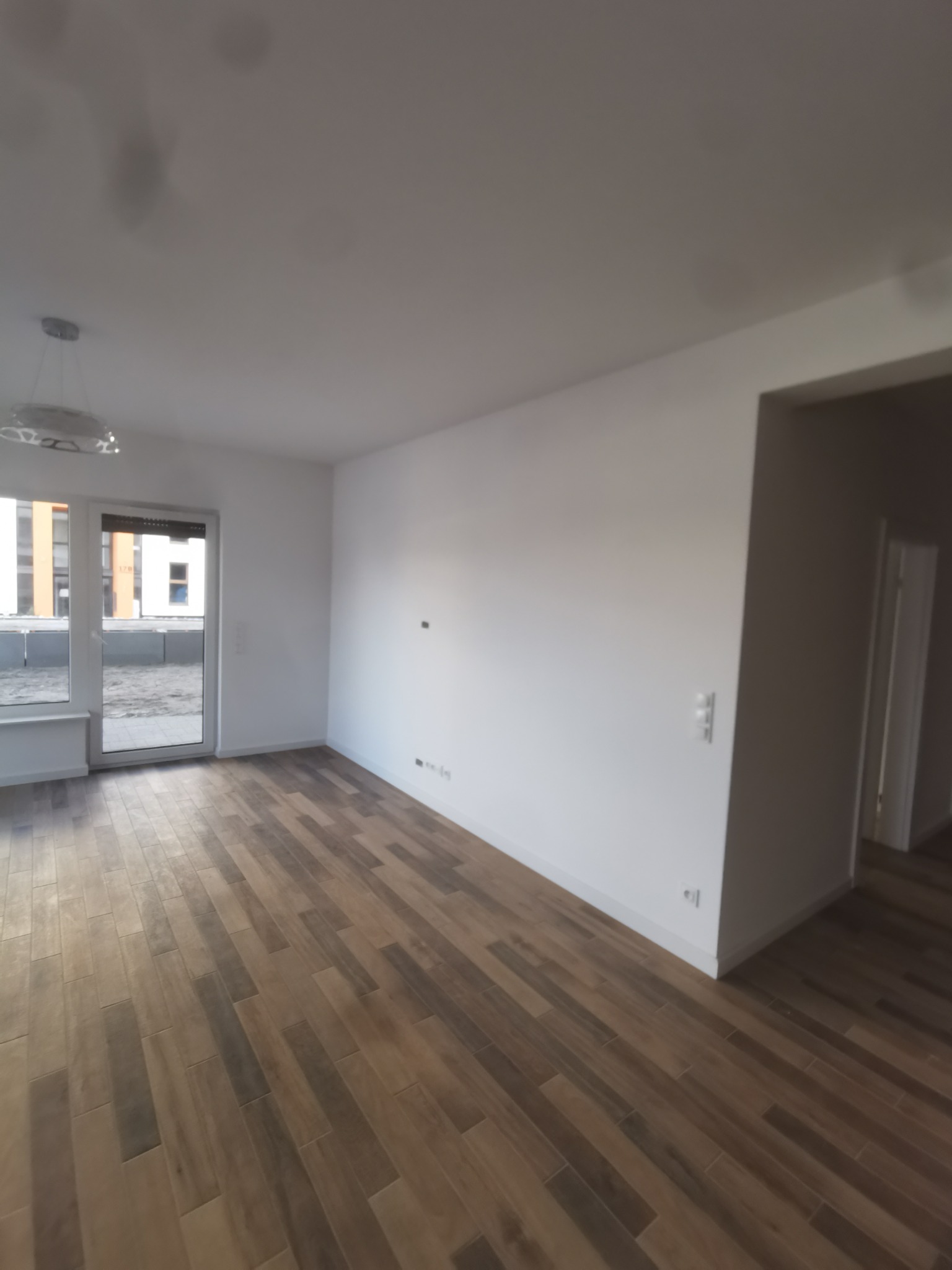 Puste, jasne pomieszczenie z panelami podłogowymi imitującymi drewno ułożonymi wzdłuż, białe ściany, okno balkonowe z widokiem na inne budynki, widoczny sufit z lampą.