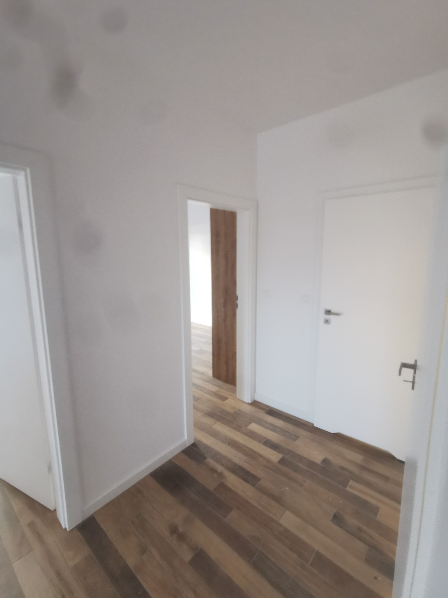 Korytarz po remoncie z drewnopodobną podłogą ułożoną w jodełkę, białe ściany i drzwi, widok na kolejne pomieszczenie z drewnianą ścianą.