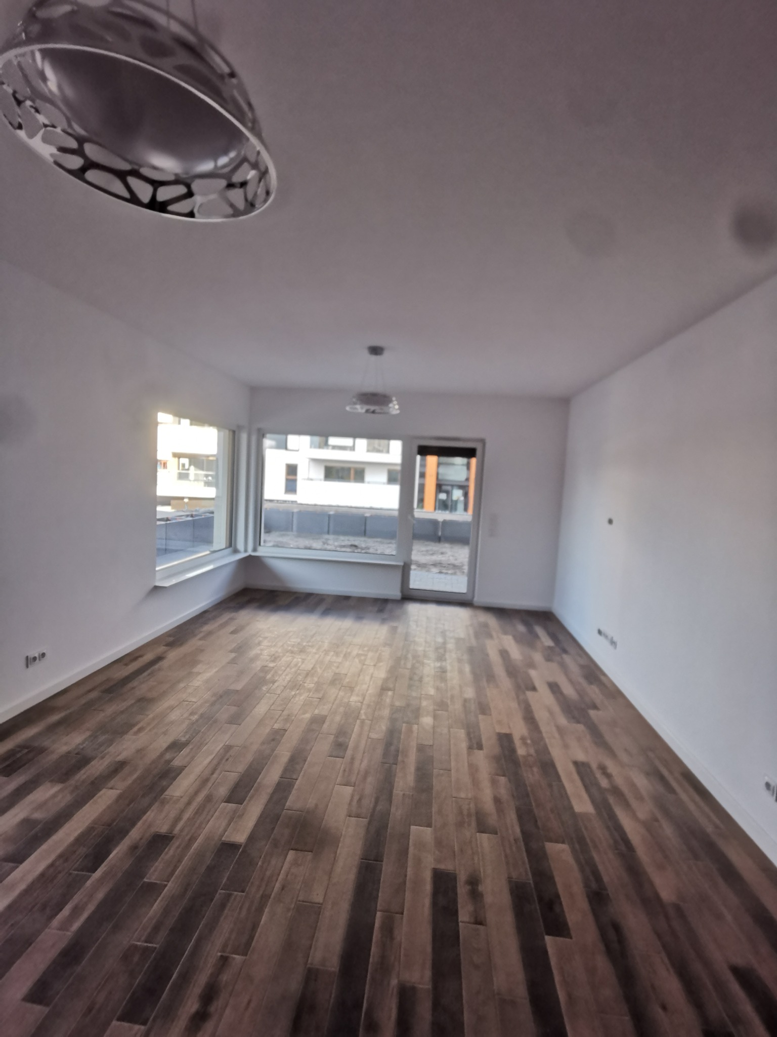 Puste, świeżo wykończone wnętrze mieszkania z panelami podłogowymi w odcieniach brązu, białymi ścianami i widokiem na osiedle za oknem.