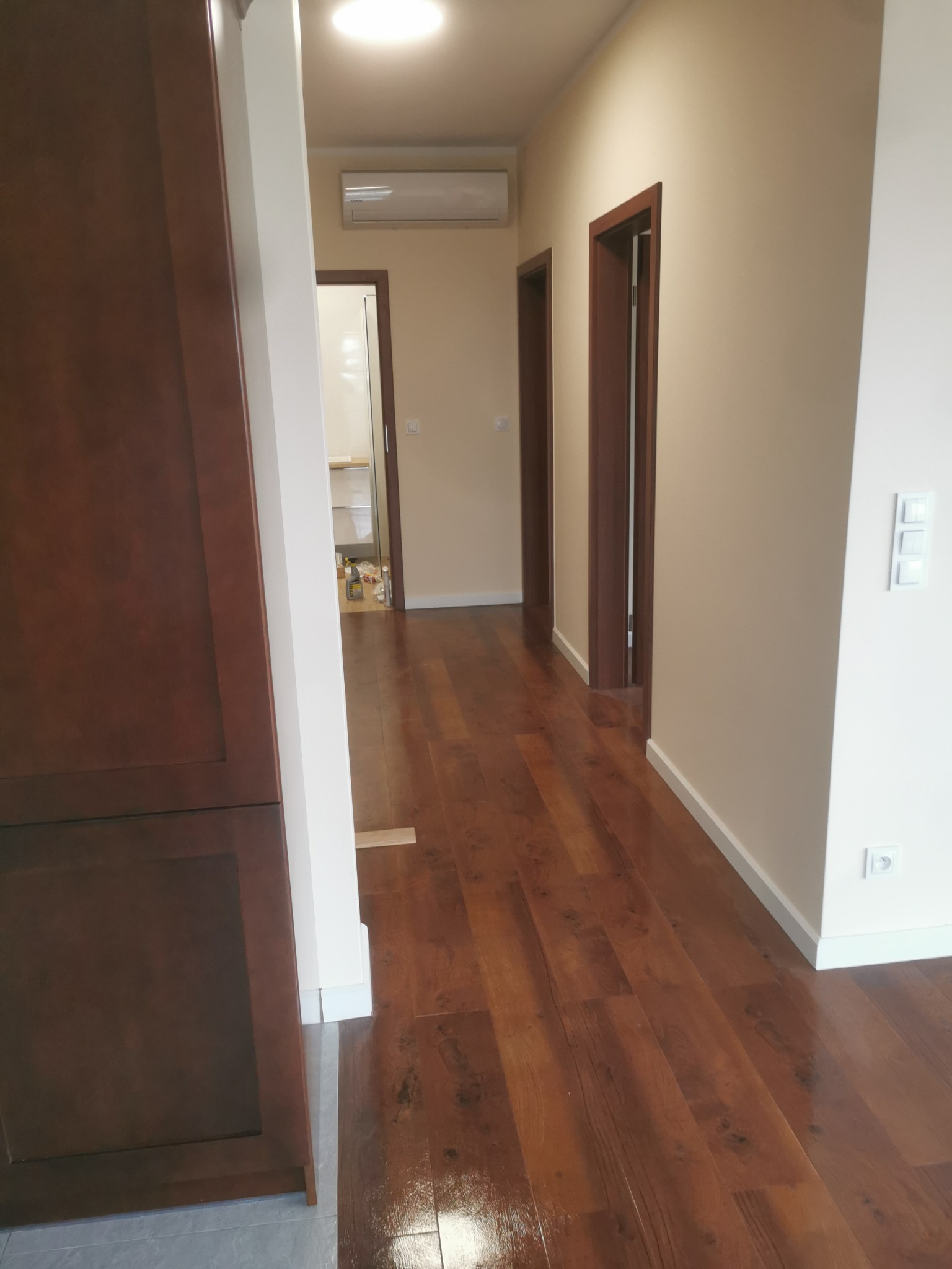 Korytarz po remoncie z nową podłogą z paneli w odcieniach brązu, widoczne drzwi z ciemnego drewna i białe ściany z zamontowanymi włącznikami.
