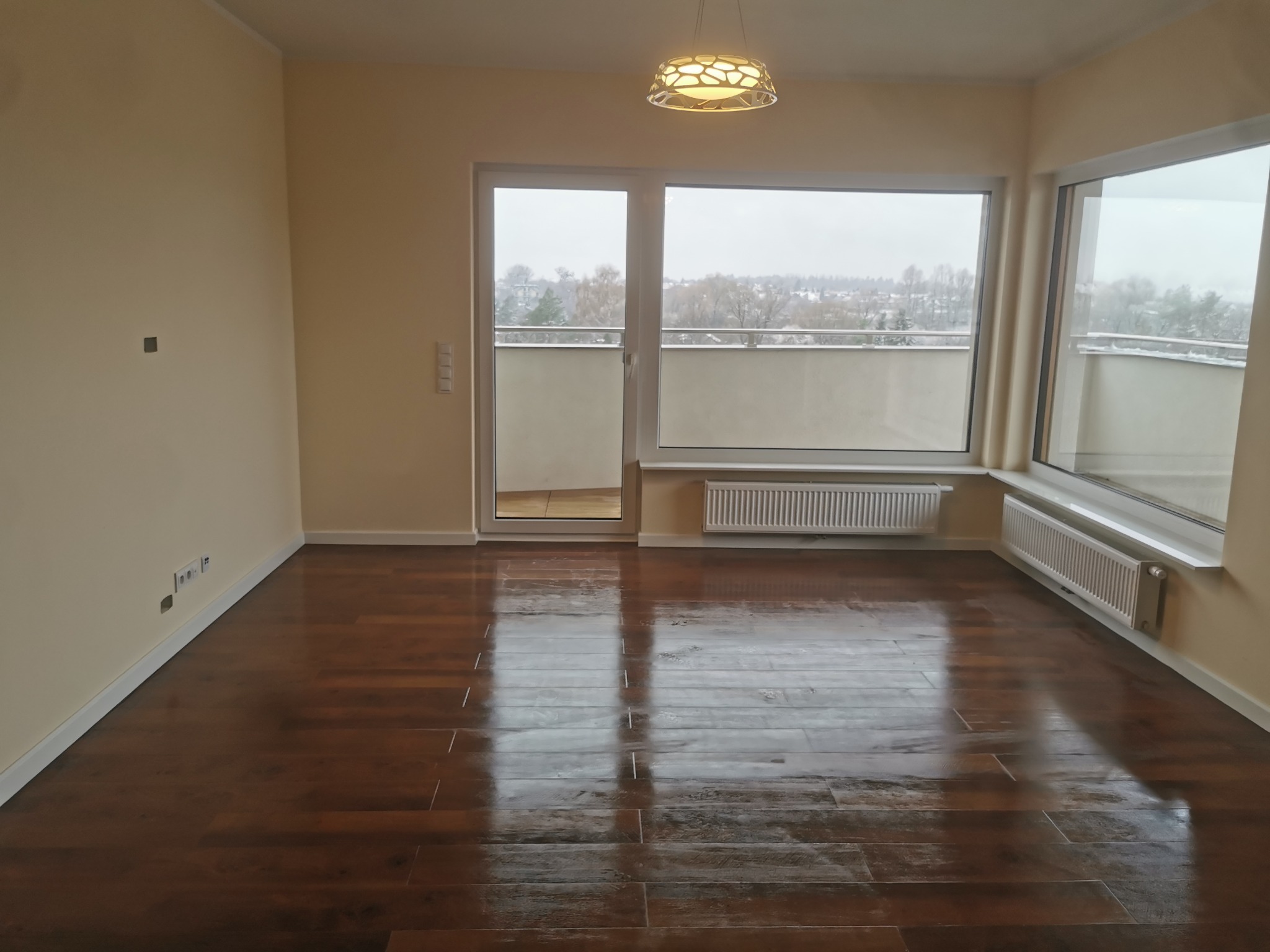 Pusty pokój w Opolu po remoncie, z parkietem ułożonym w jodełkę, widok na zaśnieżone drzewa z balkonu i okna, designerska lampa sufitowa.