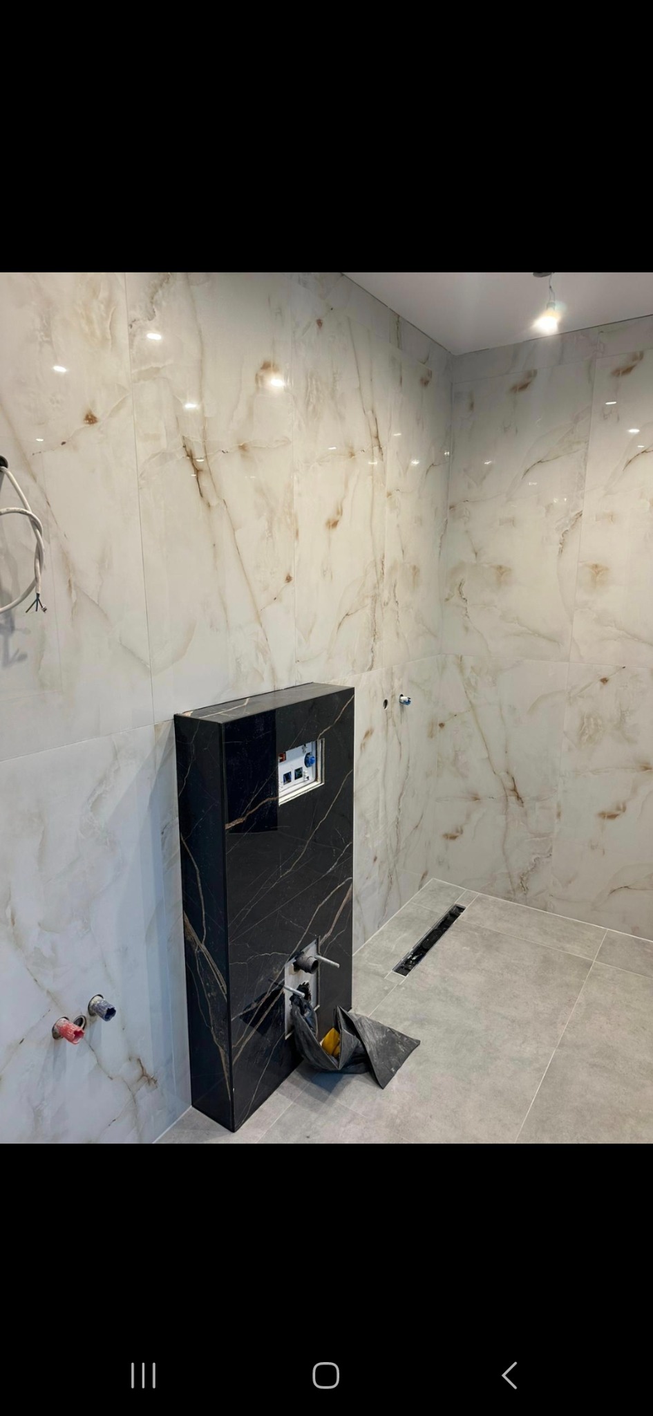 Nowoczesna łazienka w trakcie remontu: marmurowe płytki ścienne, szary gres podłogowy, czarna zabudowa stelaża WC z widocznymi przyłączami hydraulicznymi i odpływem liniowym w podłodze.