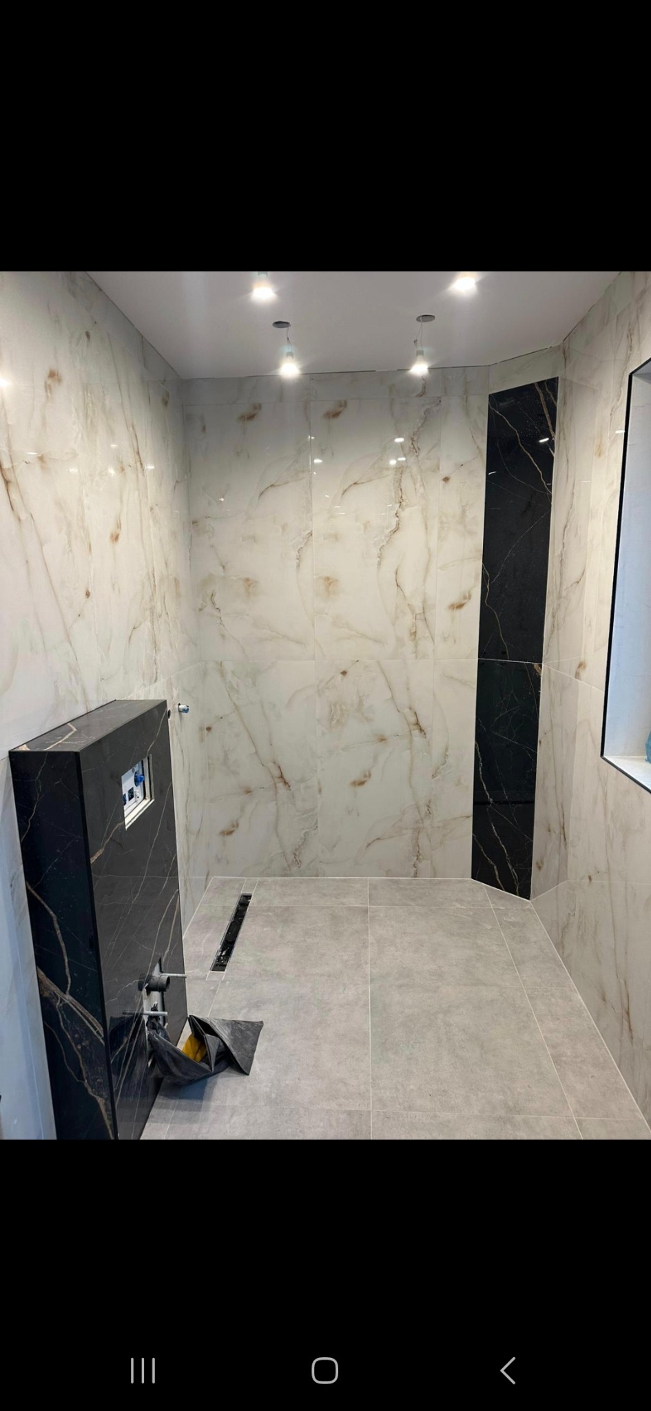 Nowoczesna łazienka z marmurowymi płytkami w odcieniach bieli i brązu, kontrastującymi z pionowym pasem czarnych marmurowych płytek; widoczny odpływ liniowy w podłodze i zabudowa stelaża WC...