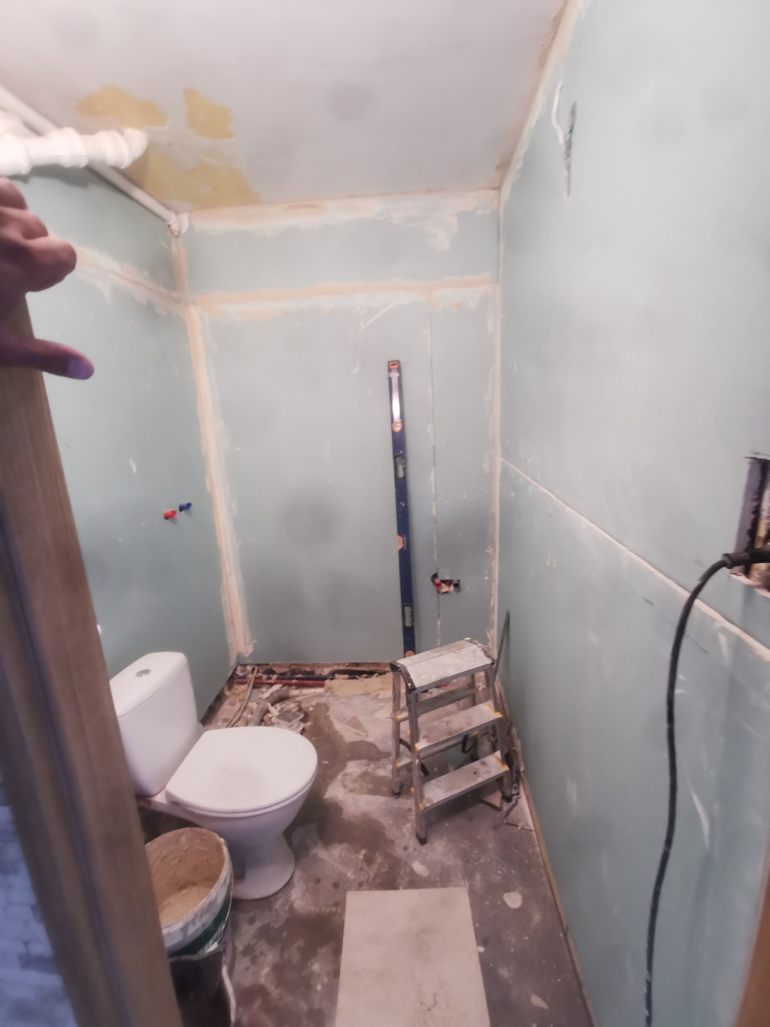 Niedokończona łazienka w trakcie prac: widoczna toaleta, poziomica oparta o ścianę z płyt karton-gips, drabinka aluminiowa, wiadro z zaprawą i fragment drzwi wejściowych.