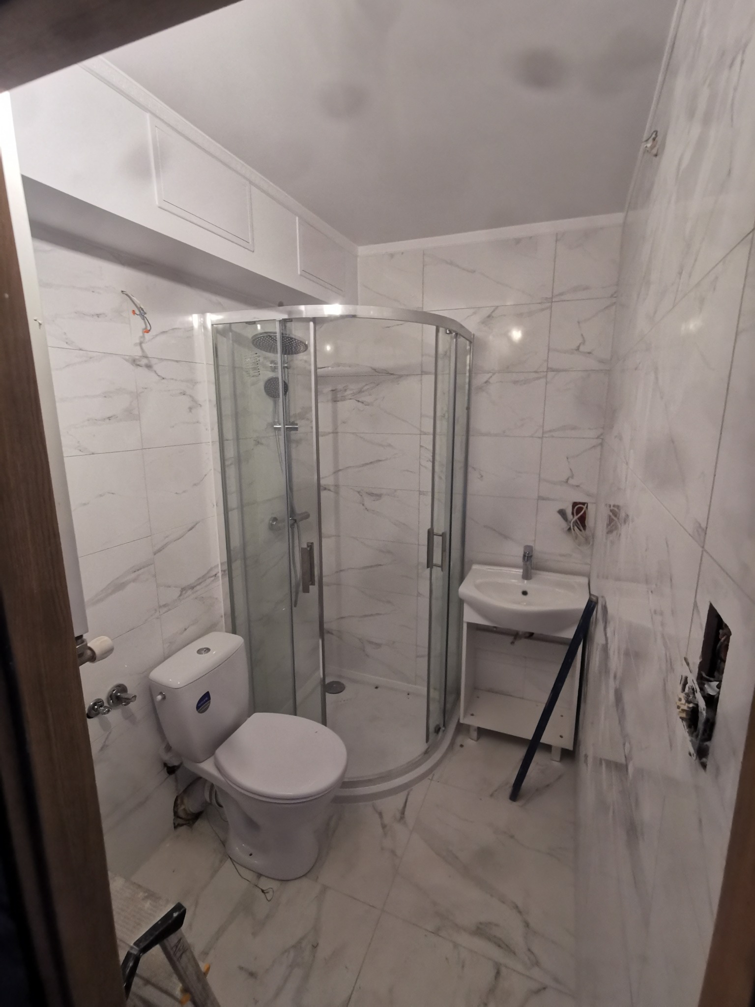 Wnętrze łazienki z kabiną prysznicową narożną, toaletą i umywalką na szafce, wykończone białymi płytkami imitującymi marmur, widoczne niedokończone prace elektryczne i drabina.