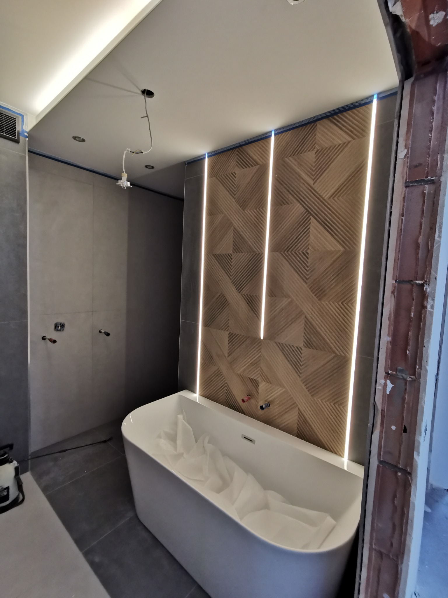 Nowoczesna łazienka w Opolu w trakcie wykańczania: wolnostojąca wanna zabezpieczona folią, szare płytki, dekoracyjna drewniana ściana z podświetleniem LED, widoczne niedokończone obrzeża.