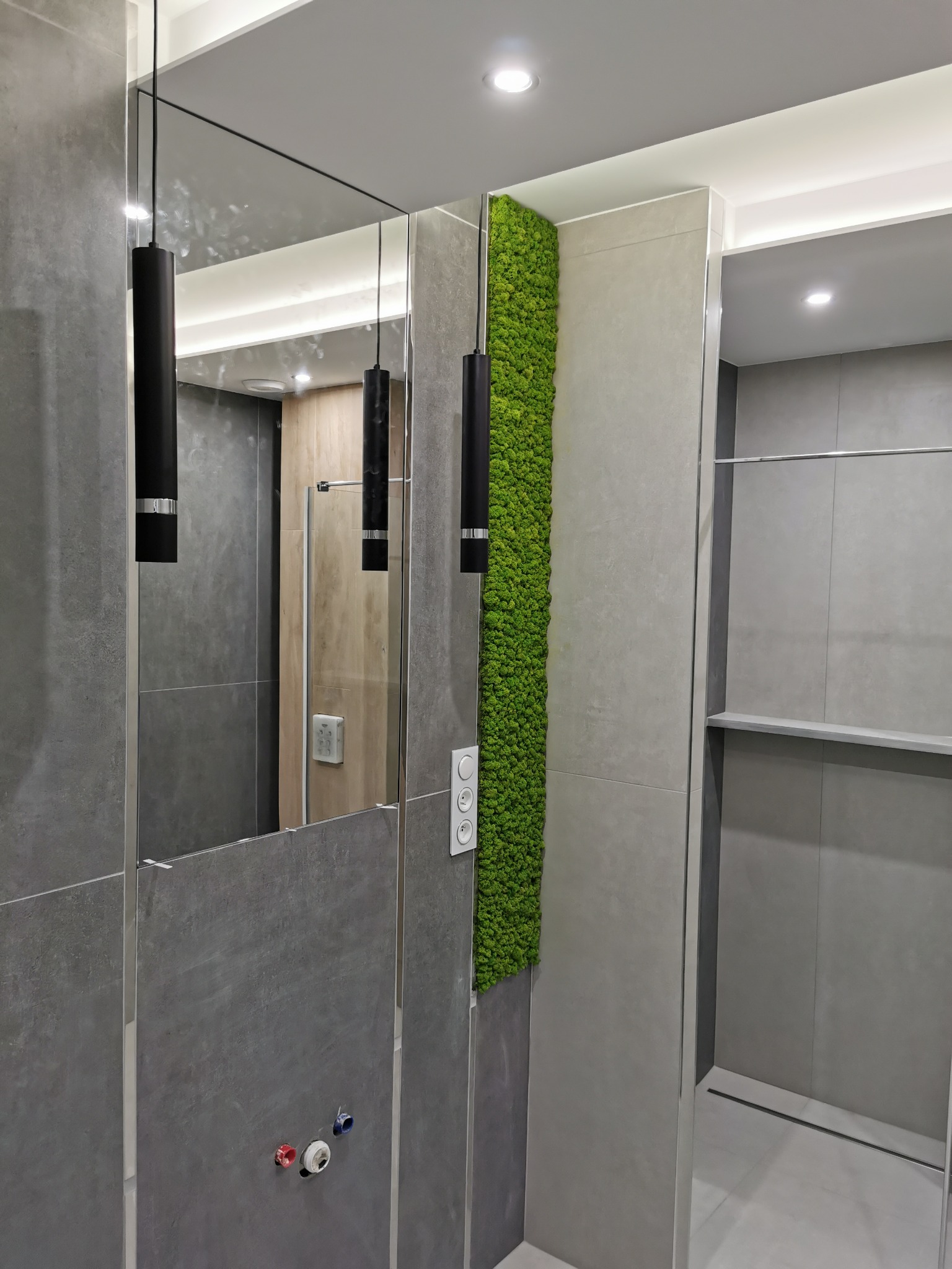Nowoczesna łazienka z szarymi płytkami, pionowym panelem z mchu, podwieszanymi czarnymi lampami i dużym lustrem.