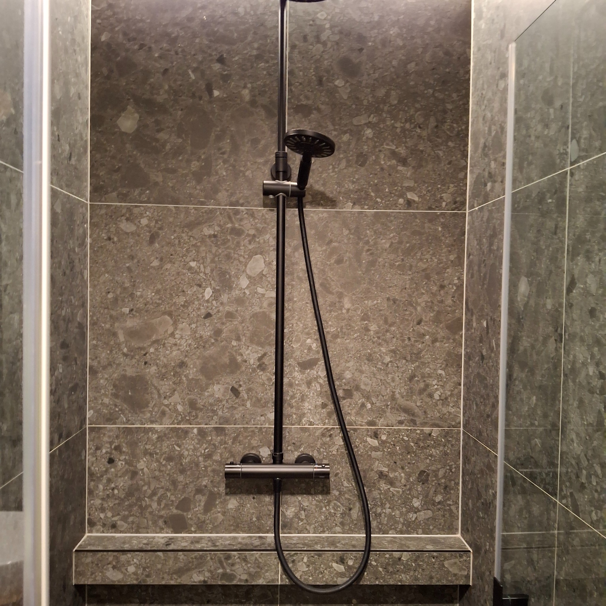 Nowoczesna kabina prysznicowa z ciemnoszarą armaturą i płytkami imitującymi kamień, widoczny fragment szklanej ścianki.