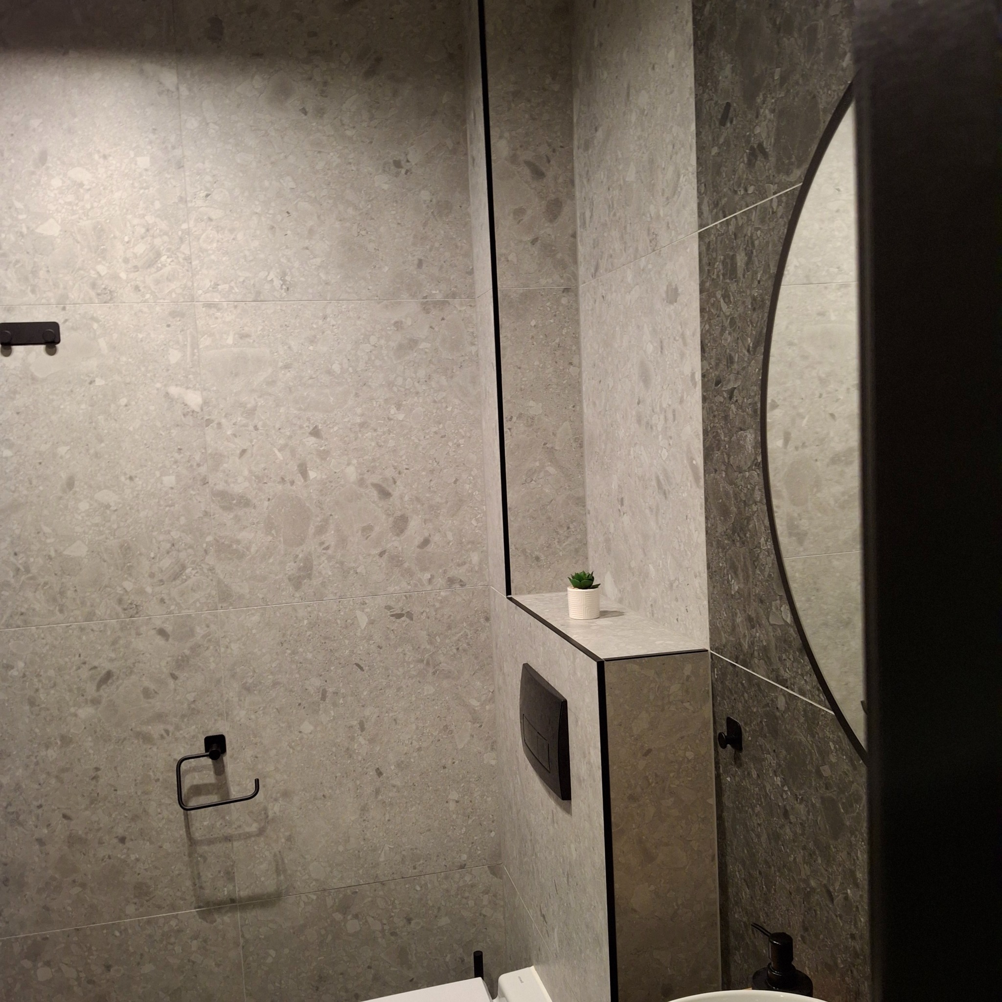 Nowoczesna łazienka z szarymi płytkami imitującymi kamień, czarnymi akcentami w postaci ramy lustra i armatury, oraz małą roślinką na półce.