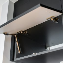 ALEKSANDRA BORAWSKA - Otwarta szafka kuchenna z mechanizmem podnoszenia, front biały z czarnym obramowaniem, widoczne zawiasy i siłownik. Minimalistyczny design.