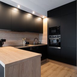 ALEKSANDRA BORAWSKA - Nowoczesna kuchnia z czarnymi, matowymi szafkami i drewnianym blatem. Oświetlenie punktowe, minimalistyczny design, drewniana podłoga. Widoczny czajnik i akcesoria kuchenne.