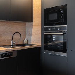ALEKSANDRA BORAWSKA - Nowoczesna, czarna kuchnia z drewnianymi akcentami: zabudowa z piekarnikiem i mikrofalówką, czarny zlew z baterią, drewniany blat i podłoga. Minimalistyczny design.