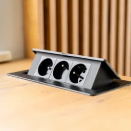 Wpuszczane gniazdo elektryczne z trzema otworami, zamontowane w blacie biurka z jasnego drewna. Widoczny fragment wtyczki w jednym z gniazd.