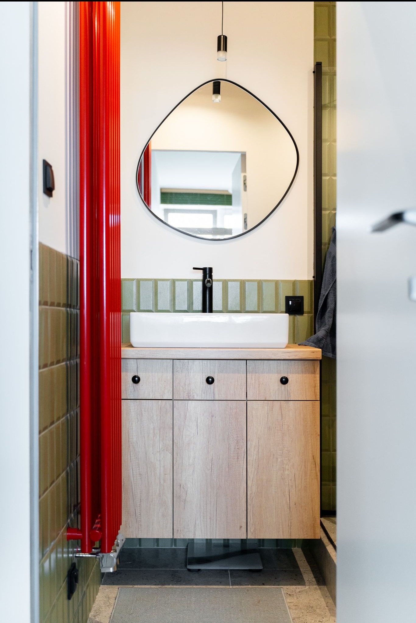 Nowoczesna łazienka z umywalką nablatową na szafce z drewnopodobnym frontem, asymetrycznym lustrem i intensywnie czerwonym grzejnikiem żeberkowym.