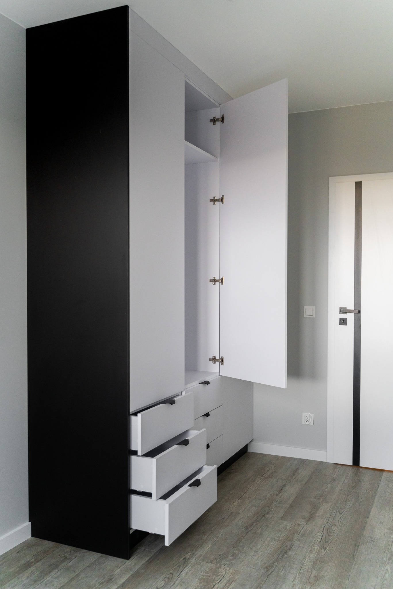Nowoczesna szafa na wymiar z czarnym frontem i otwartymi szufladami w jasnym wnętrzu. Otwarta szafa ukazuje półki. Widoczna podłoga z paneli.