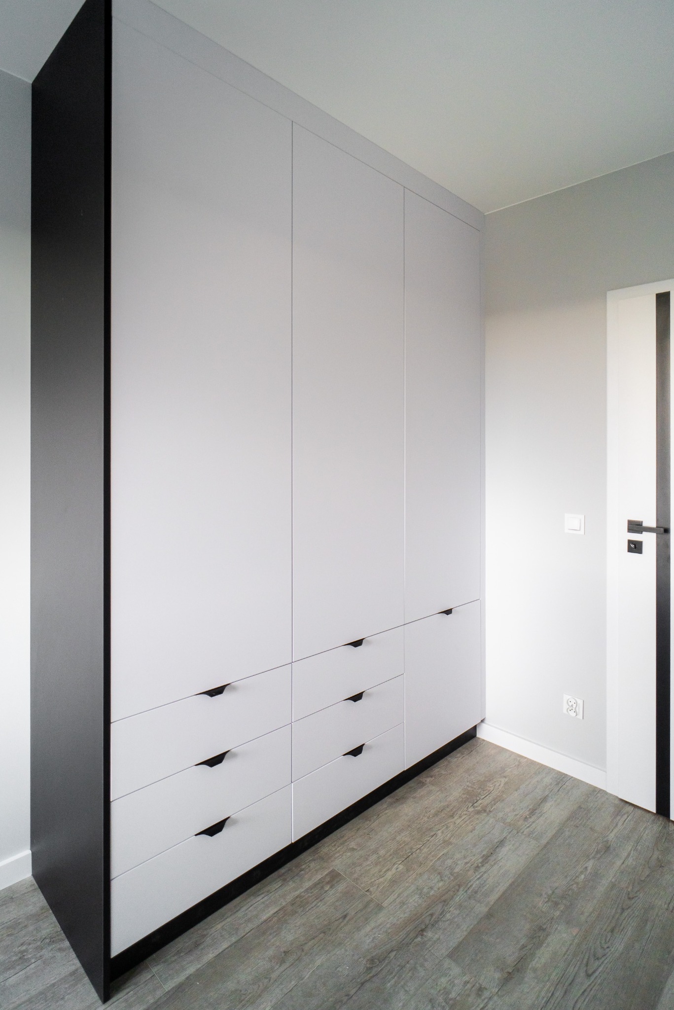 Biało-szara szafa wnękowa z czarnymi akcentami, widok z dołu. Minimalistyczny design, drewniana podłoga, szare ściany, czarne uchwyty. Nowoczesne wnętrze.